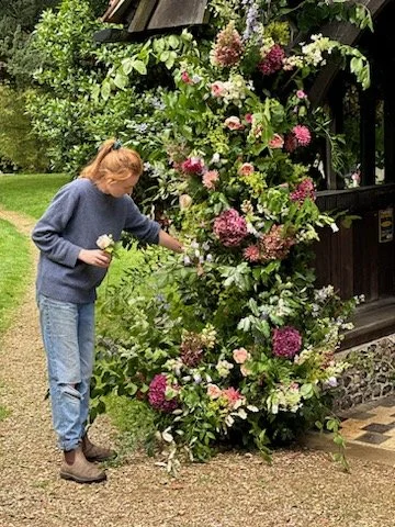 A woman watering flowers beside a large, colorful flower bush outside a building.