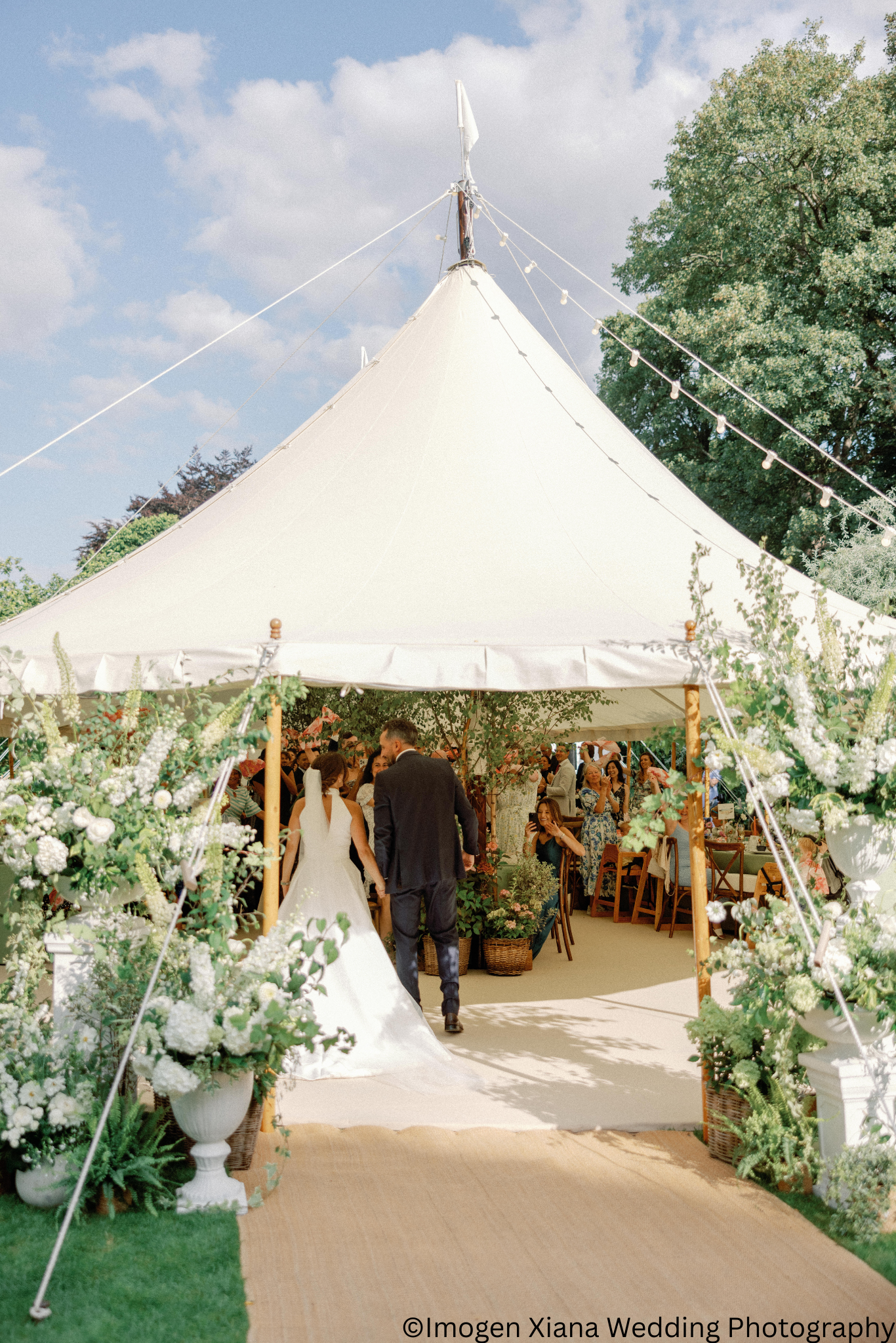 A bride and groom walk into a wedding reception tent decorated with white flowers and greenery, with guests inside under a bright sky.