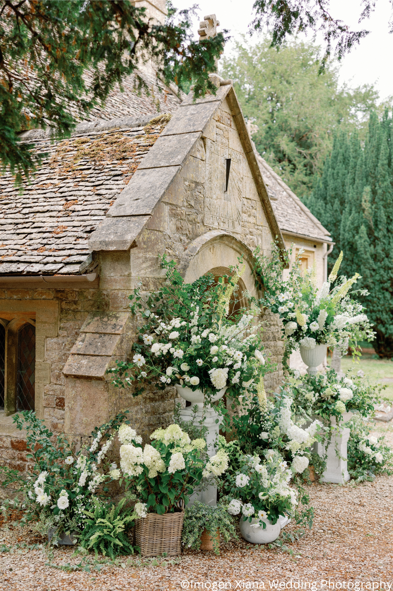 A stone church with a clock on the front, surrounded by white flowers in large vases and pots.