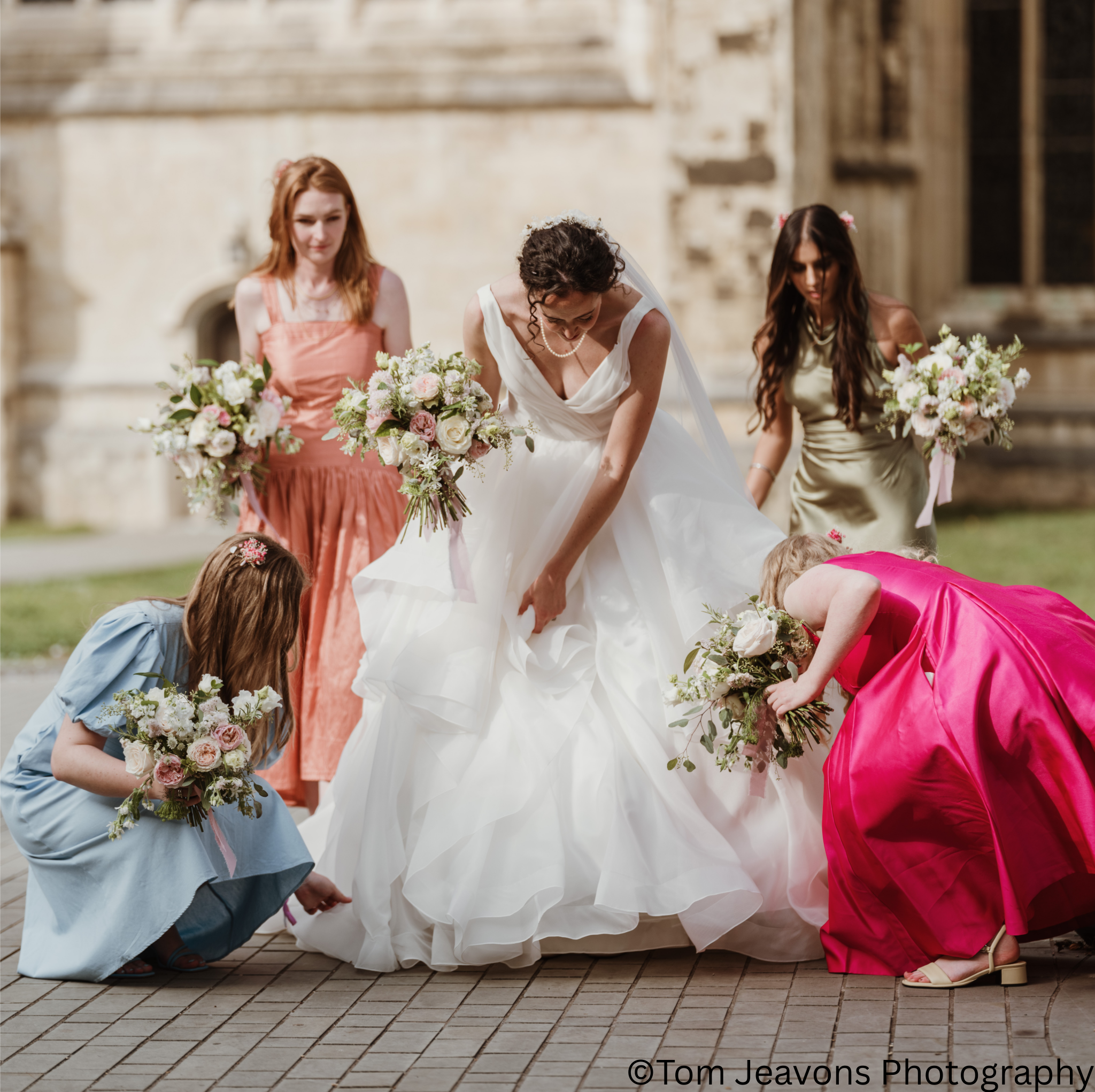 A bride in a white wedding gown is surrounded by five girls in colorful dresses, all holding bouquets, outdoors in front of a historic building.