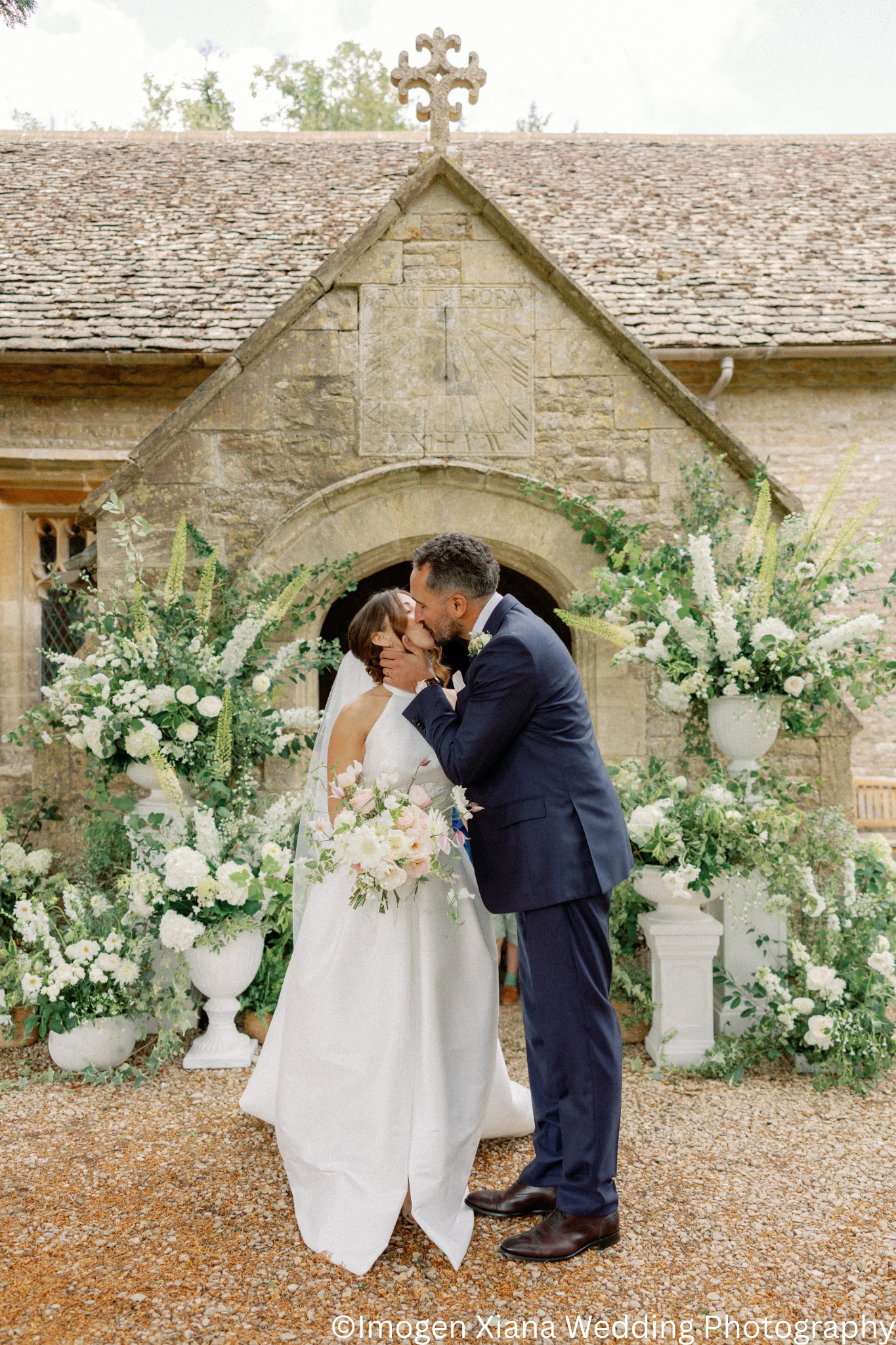 A bride and groom kiss during their wedding ceremony in front of a Cotswolds stone church.  They are surrounded by beautiful large white floral arrangements on pedestals in groupings.