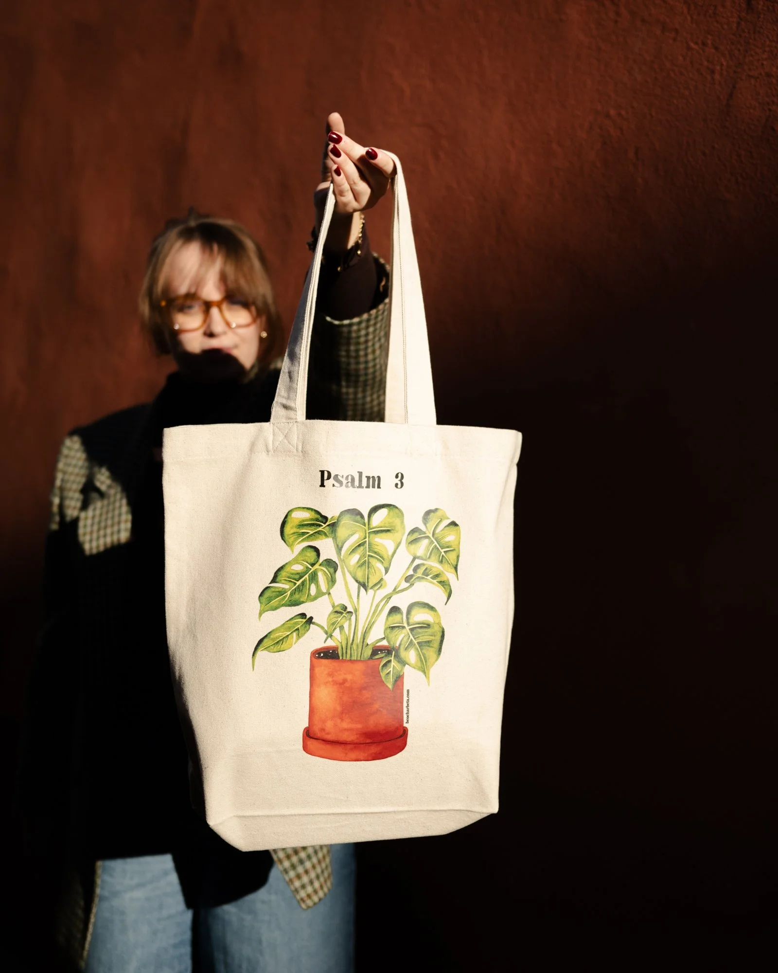 Person håller upp en vit tygväska med en illustration av en växt i en kruka med en Monstera i och texten "Psalm 3".