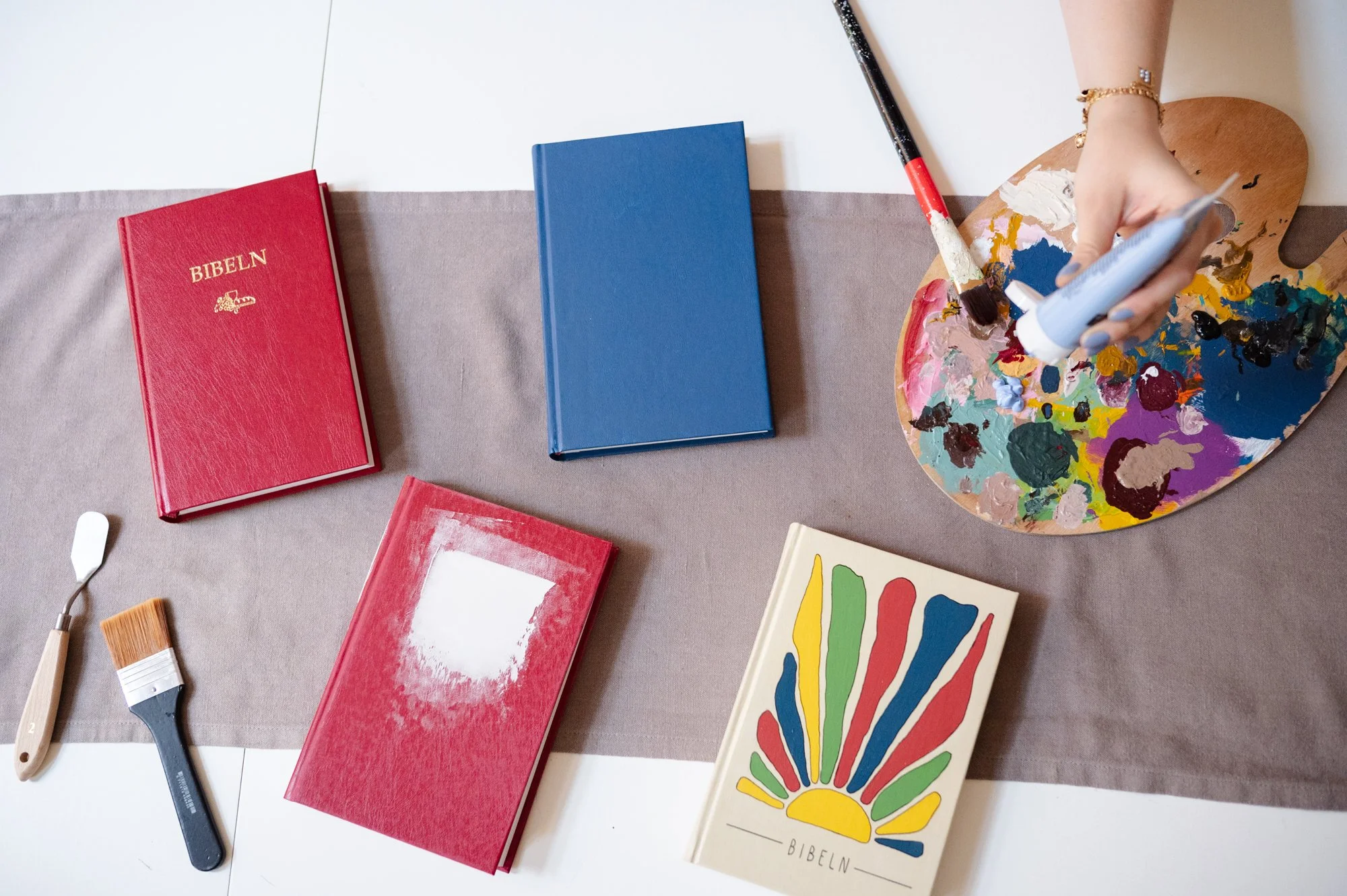 Flera Biblar av olika delar av processen till en egendesignad Bibel. En original, en slipad, en grundmålad och en färdig. Ligger på ett bord tillsammans med färg, penslar och en liten spatula.