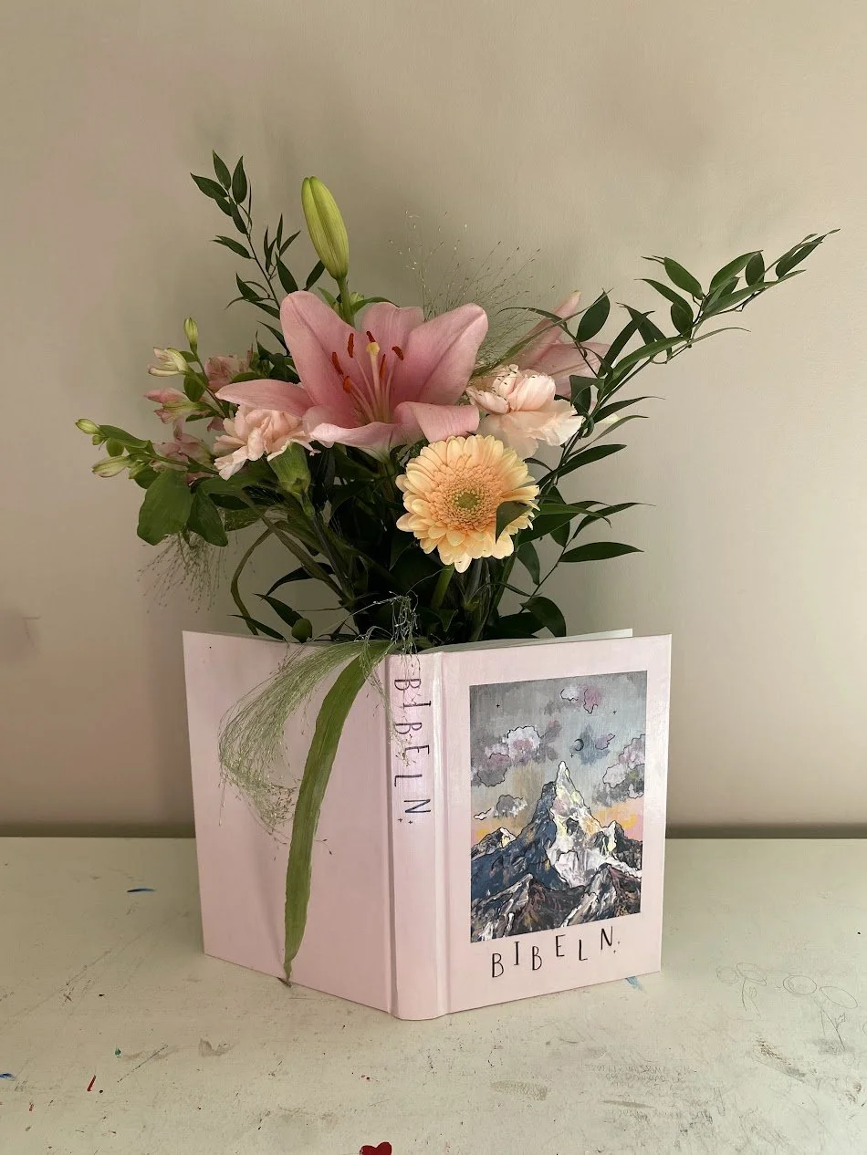 En blomsterbukett med rosa liljor, en gul gerbera och andra blommor i en bukett, placerad i en bok som är dekorerad med ett bergsmotiv och texten 'BIBELN' på dess omslag.