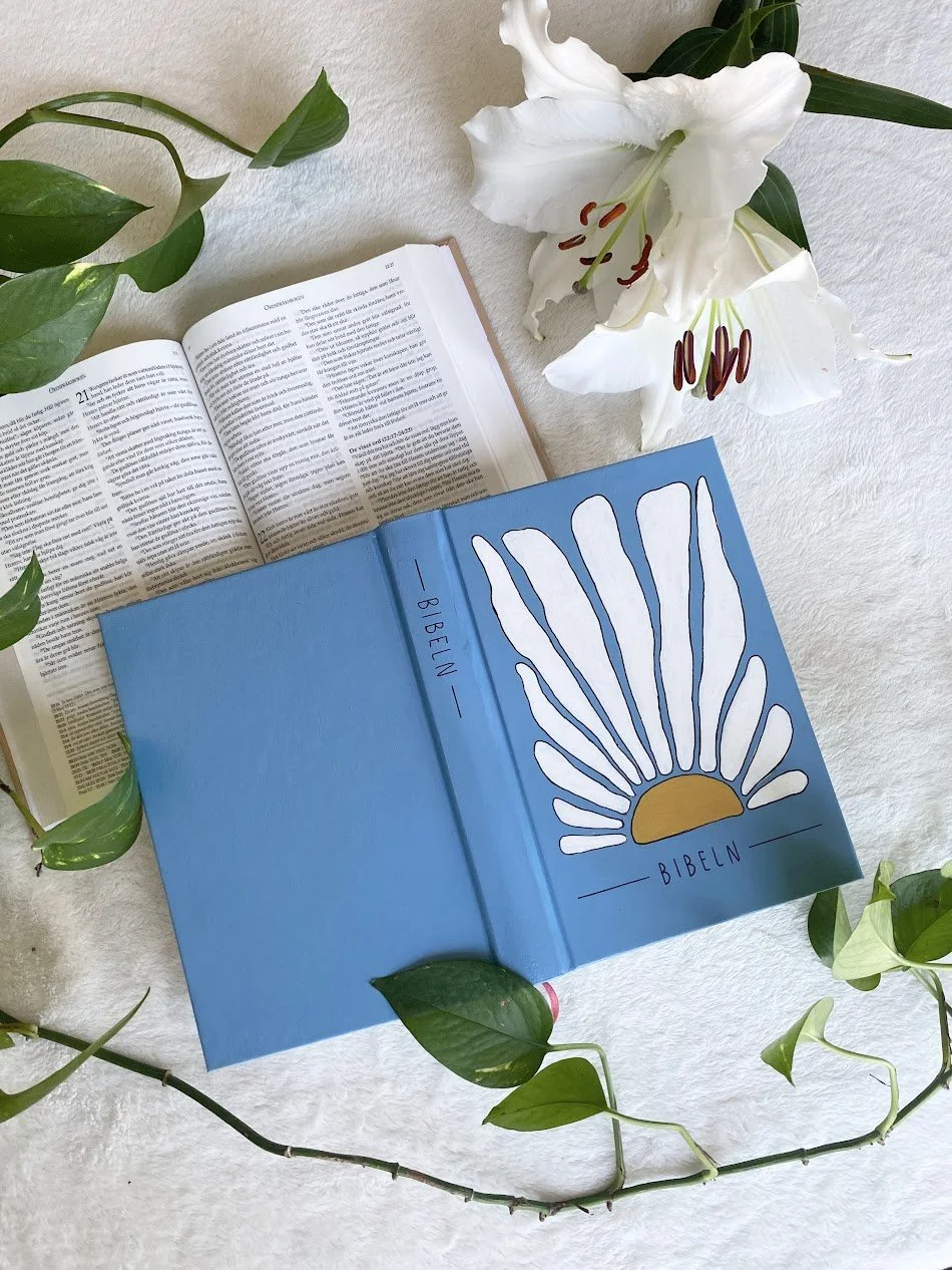 En öppen bibel, en blomsterbukett med stora vita liljor och gröna blad, placerad på en ljus textilbakgrund.