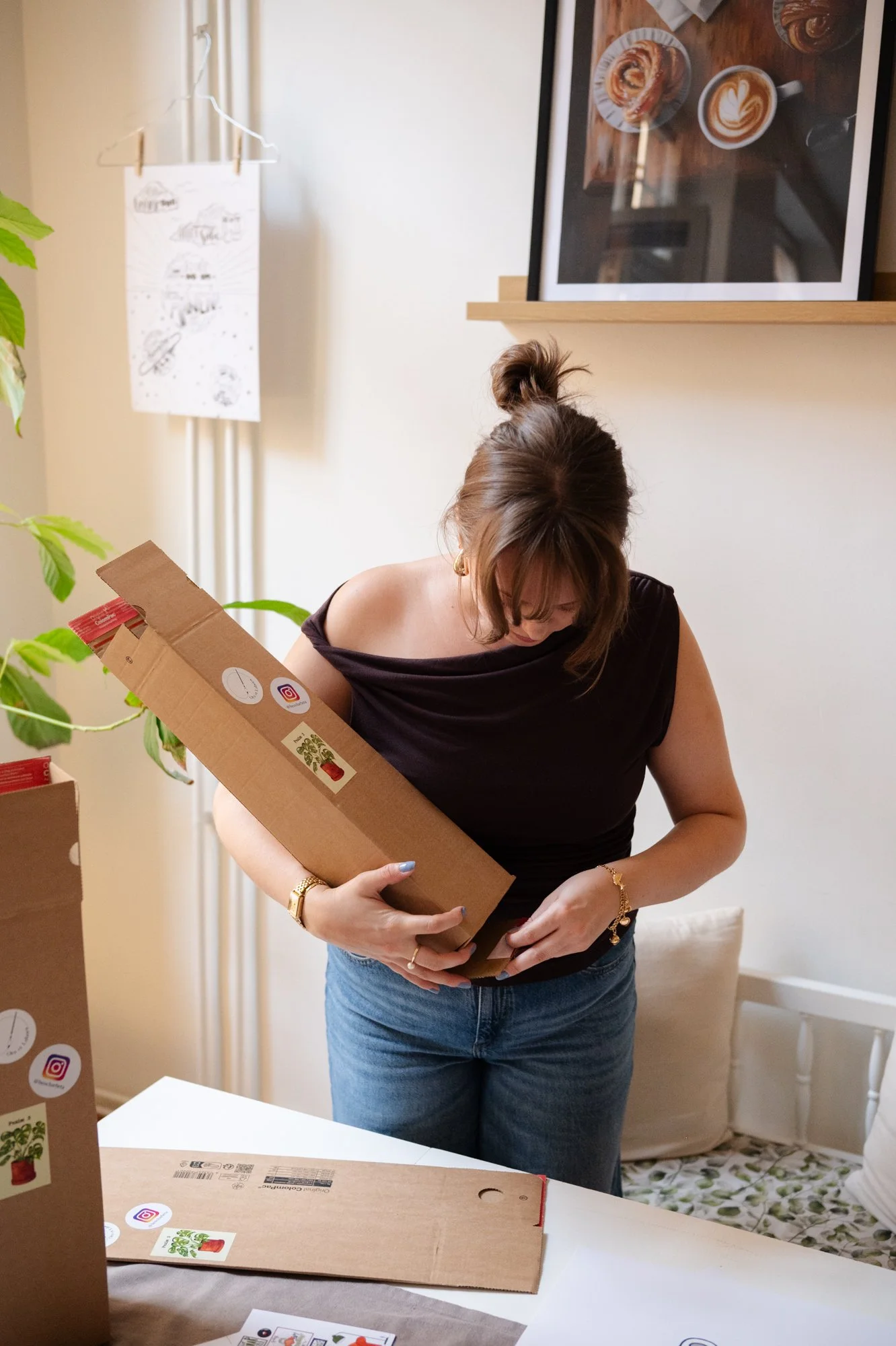 Kvinna håller i en kartong i ett rum med gröna växter och väggdekorationer.