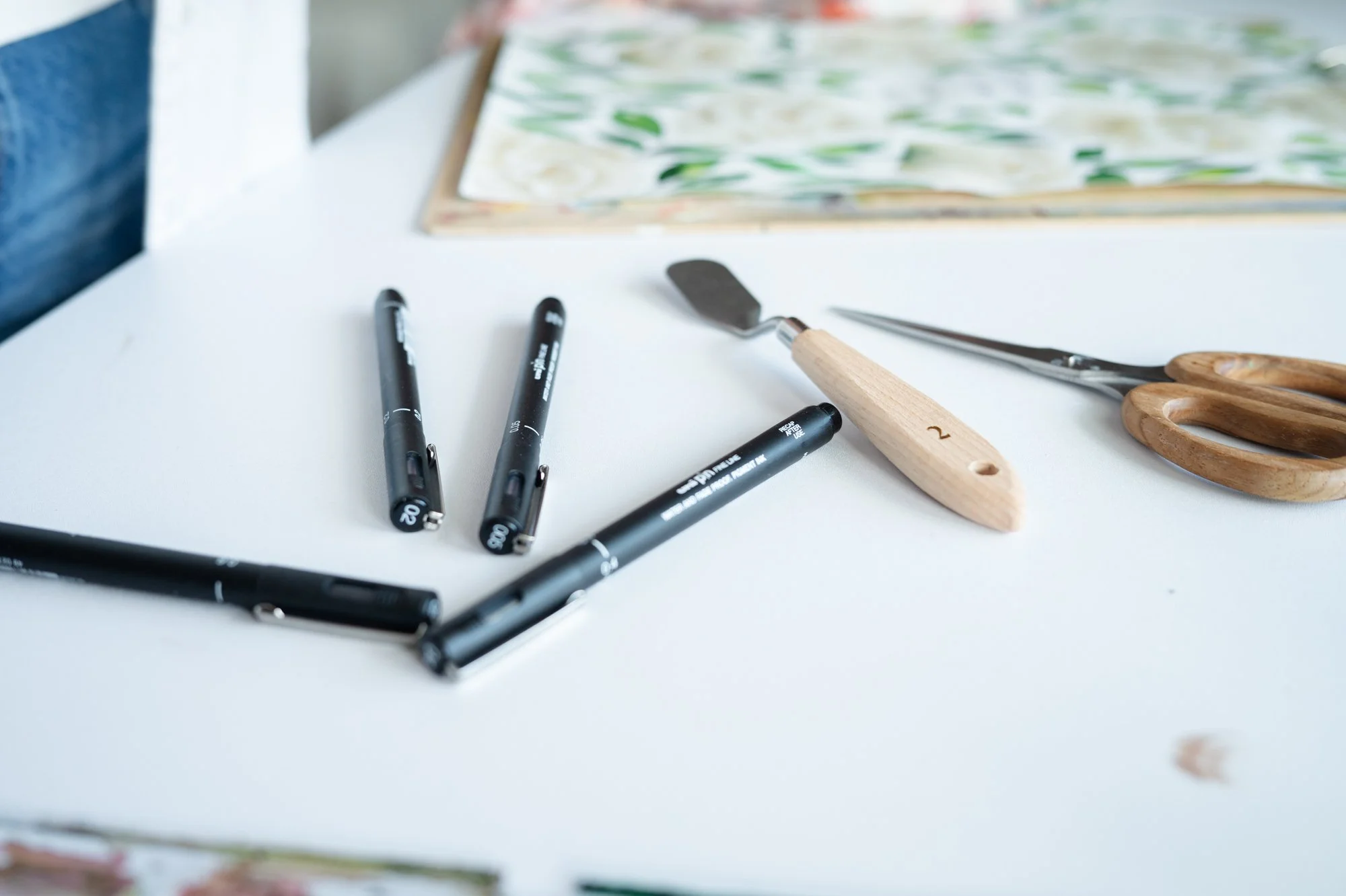 Pennor, skrapa, pincett och sax på en vit bordsyta med konstnärsmaterial i bakgrunden.