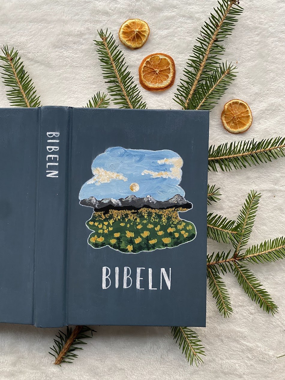 En blå bibel med illustration av landskap, moln, sol och blommor på omslaget, omgiven av granbarr och torkade apelsinskivor på en vit bakgrund.