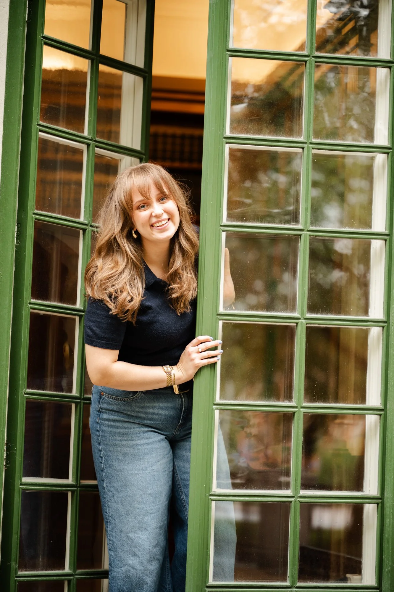 En glad ung kvinna med brunt, lockigt hår som står i en öppen glasdörr med gröna spröjs.
