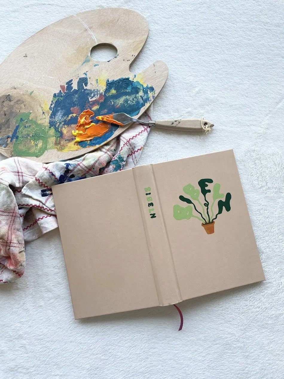 En målarduk, en palett med färg och en palettekniv ligger på ett vitt tyg tillsammans med en öppen bok som visar en illustration av en växt i en kruka.