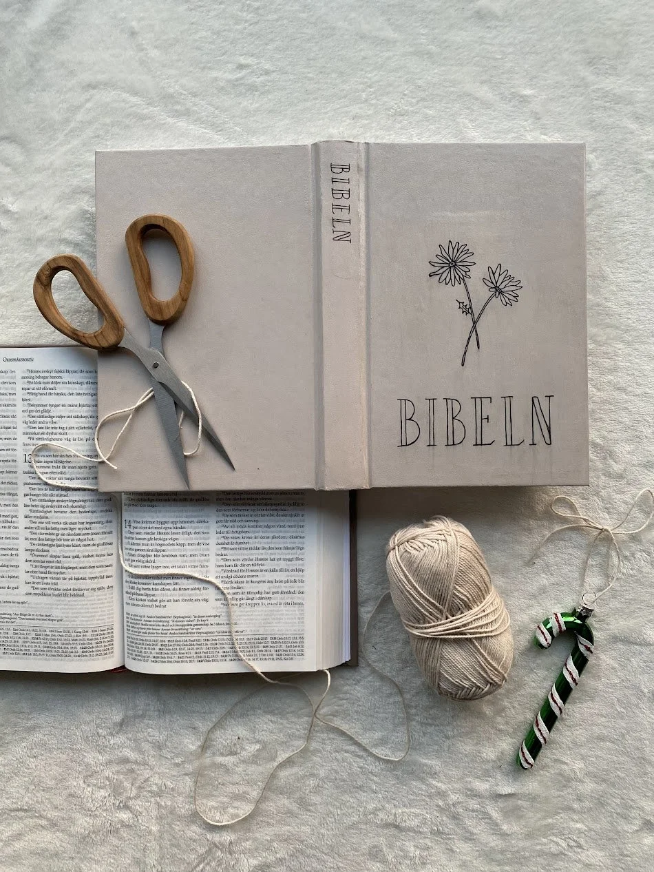 En öppen bibel, ett snittpar av sax, ett nystan av garn, en julstrumpa dekorerad med godisränder, och ett vitt anteckningsblock med texten 'BIBELN' och en blomma, ligger på en vit texturerad yta.