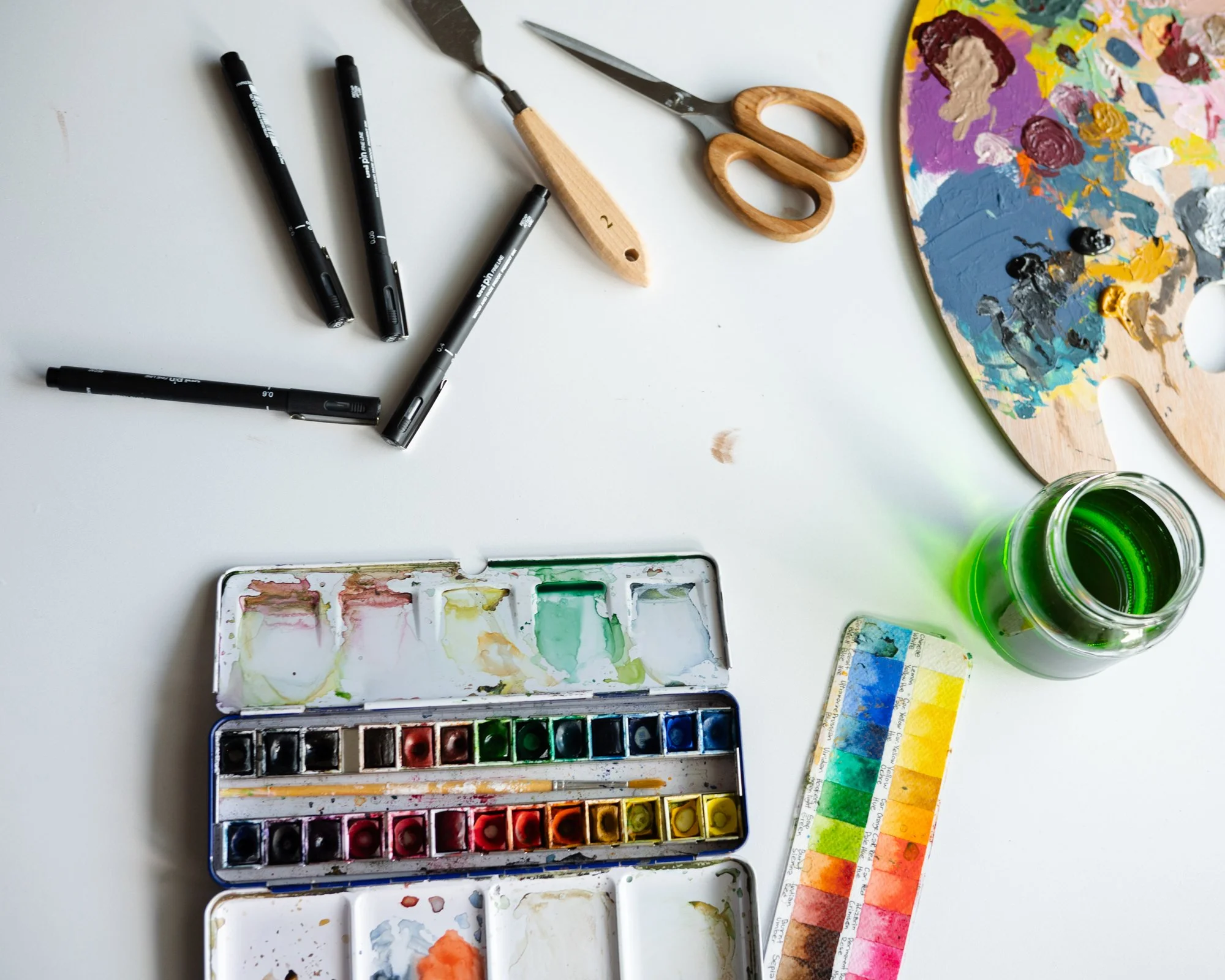 Akvarellfärger i en låda, penslar, sax, och färgprover på ett konstnärsjobb.