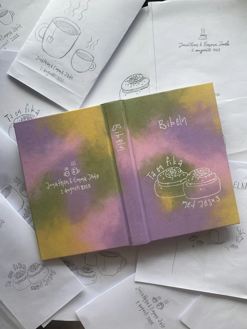 Färgad bok med titeln Bibel och illustration av kanelbullar och texten Ta en fika, samt handritat omslag för en bibel. Runt, vitt papper med handritade skisser av muggar, kaffekoppar, kanelbullar och text, med namn och datum.
