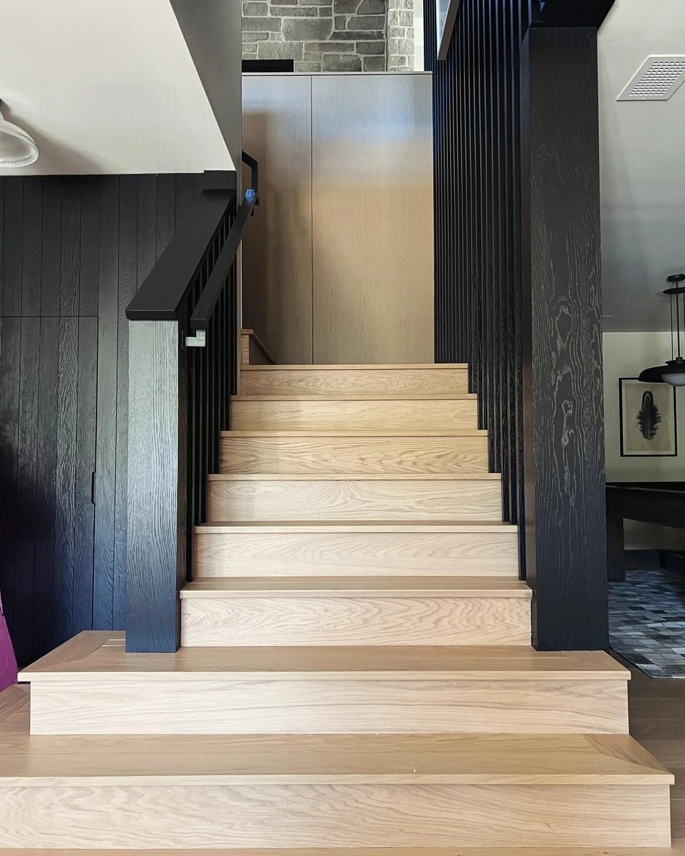 Interior view of a staircase with light wooden steps, black handrails, black vertical slats on the sides, and a partial view of a living room with framed artwork and a chandelier in the background.