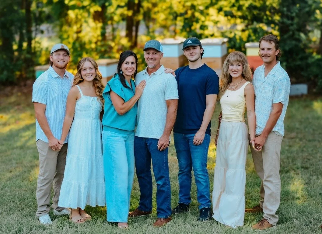 A family of seven standing outdoors on grass, with trees and beehives in the background. They are smiling and dressed casually for warm weather.