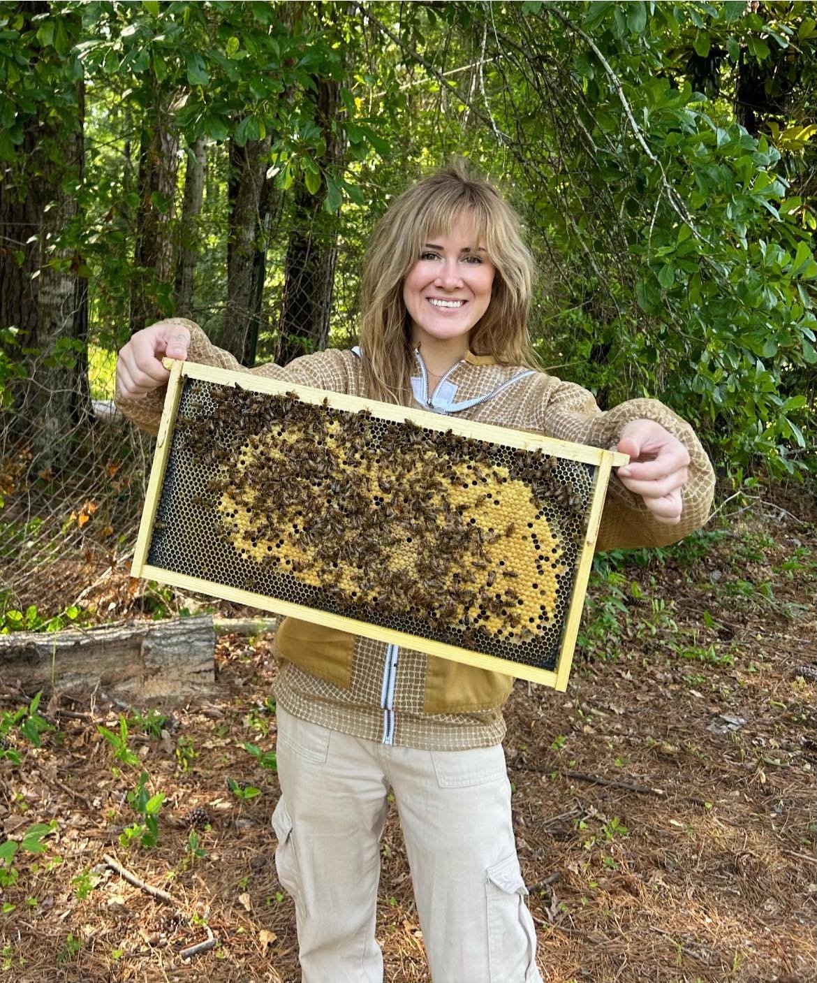 Woman holding a beekeeping frame with bees, outdoors in a wooded area.