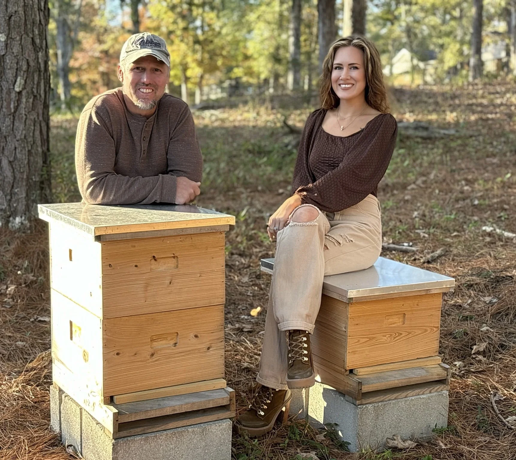A man and a woman sitting outdoors in a wooded area with autumn foliage. The man is leaning on a wooden box hive and the woman is sitting on another similar hive, both smiling.