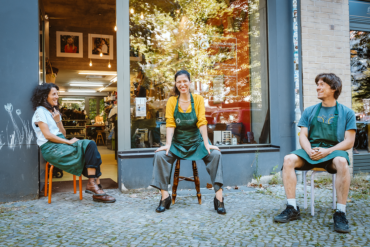 Drei Personen sitzen vor einem Geschäft einer Schuhmacherei in Berlin auf der Straße und lachen. Es wird gerade ein neues Teamfoto von der Hauptstadtlinse Fotografin Nadine Domke gemacht