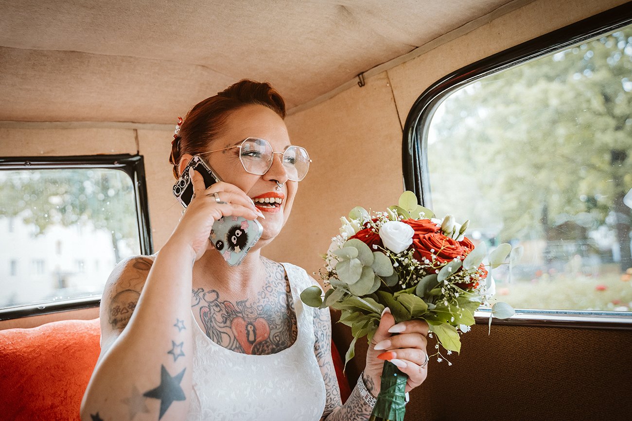 Eine Frau mit Tattoos, Brille und roten Haaren hält einen Blumenstrauß und spricht lächelnd am Handy, sitzend in einem Fahrzeug. Fotoshoot mit der Hauptstadtlinse nach der Trauung im Rathaus Pankow