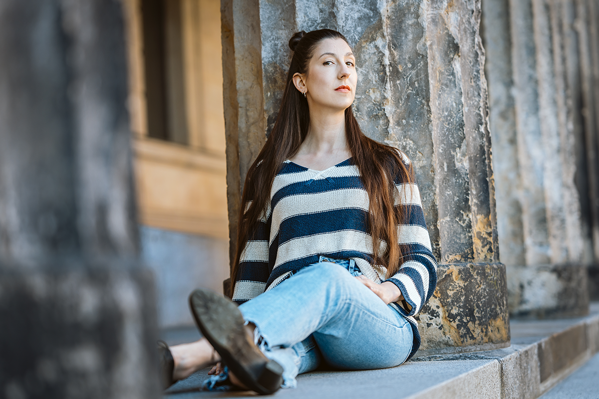 Junge Frau sitzt an einer alten Steinsäule im Freien, trägt einen gestreiften Pullover und Jeans, schaut nach oben. Sie ist Schauspielerin und heisst Rahel Schöppenthau