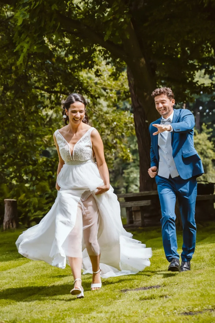 Ein lachendes Brautpaar läuft in einem Park mit grünen Bäumen und Wiese. Die Braut trägt ein weißes Hochzeitskleid und offene Schuhe, der Bräutigam trägt einen blauen Anzug.