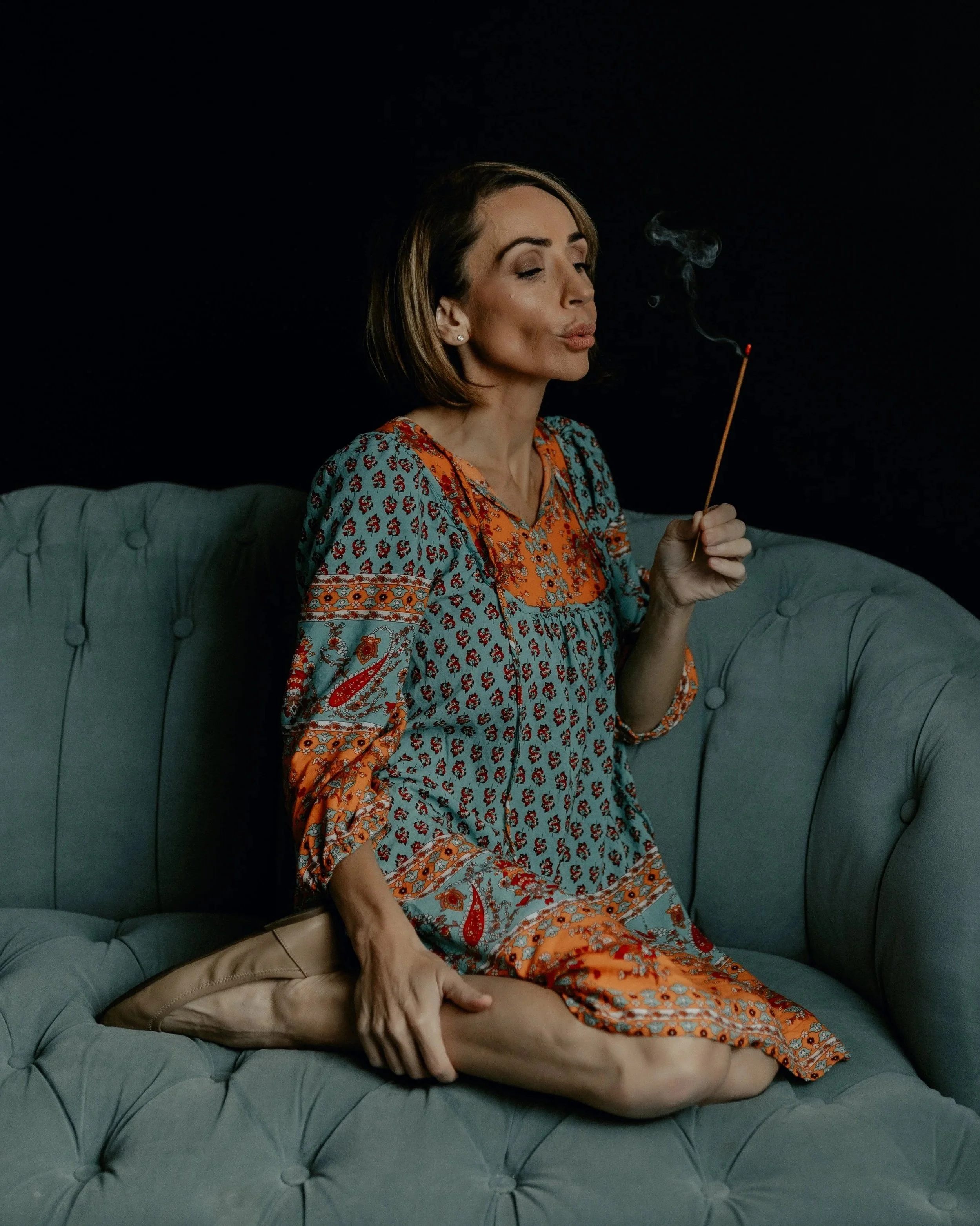 A woman with short brown hair sitting on a green tufted couch, wearing a colorful patterned dress, holding a lit matchstick, and blowing smoke.
