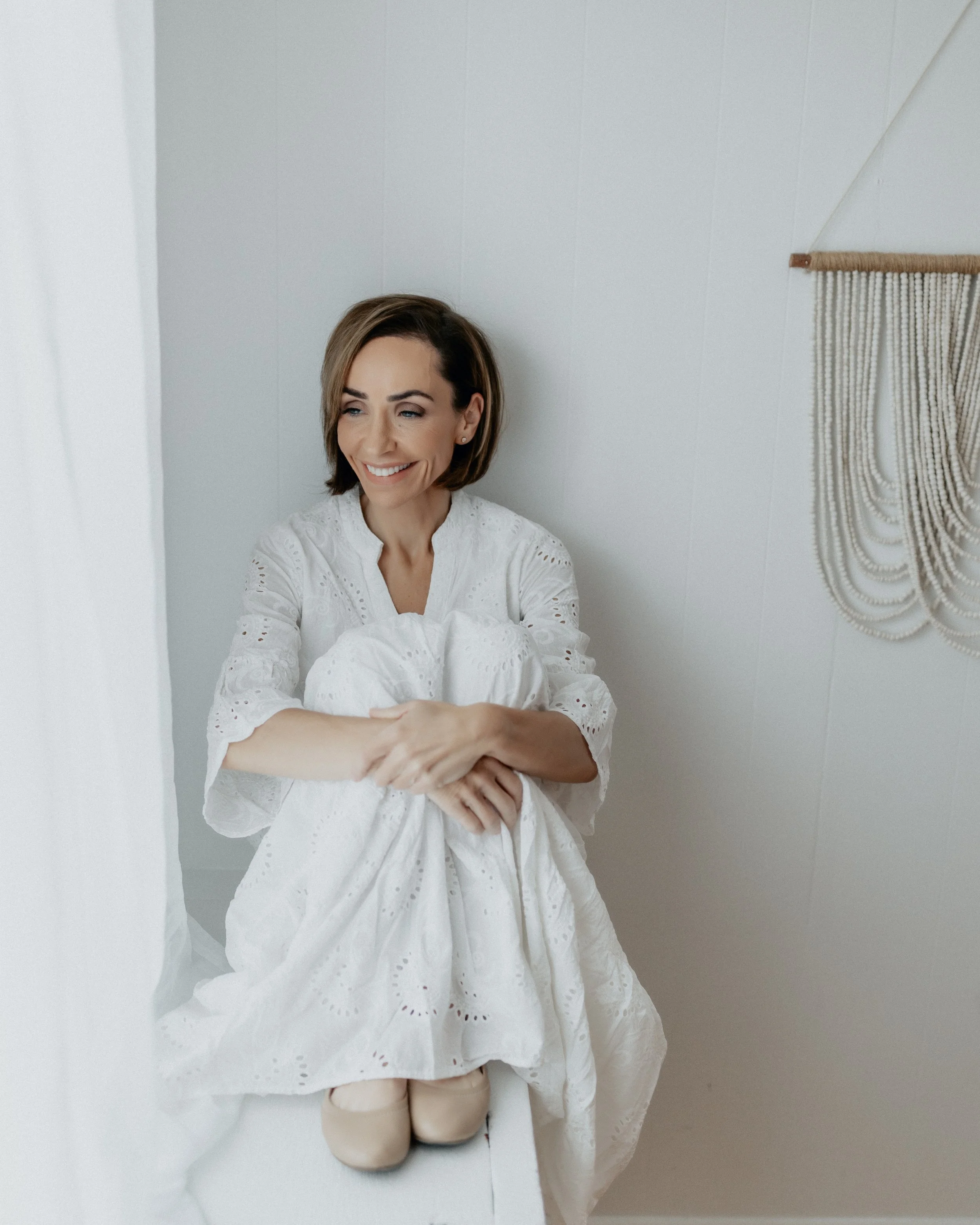 A woman with short brown hair, wearing a white dress, sitting on a white surface near a white wall, smiling and looking to the side.