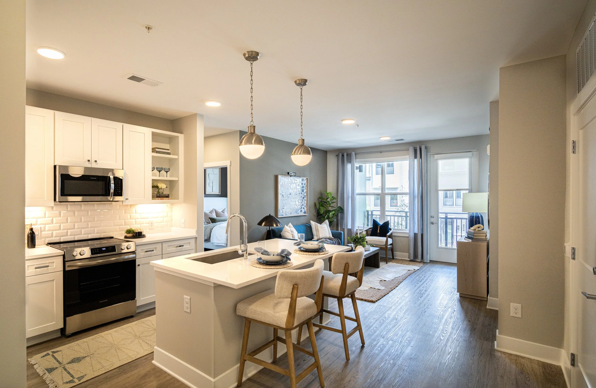 Open-concept living room and kitchen with white cabinets, stainless steel appliances, a kitchen island with barstools, and a seating area with couch, chair, and windows letting in natural light.