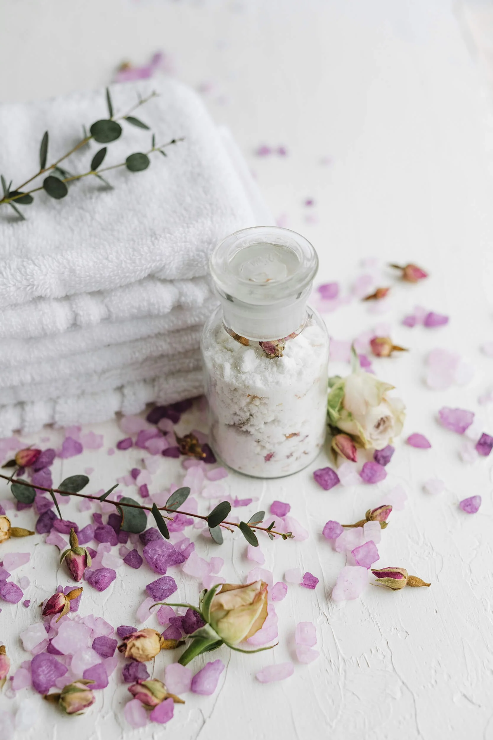 A bottle of bath salts, white towels, and dried pink roses representing massage therapy in Chesterfield, VA..