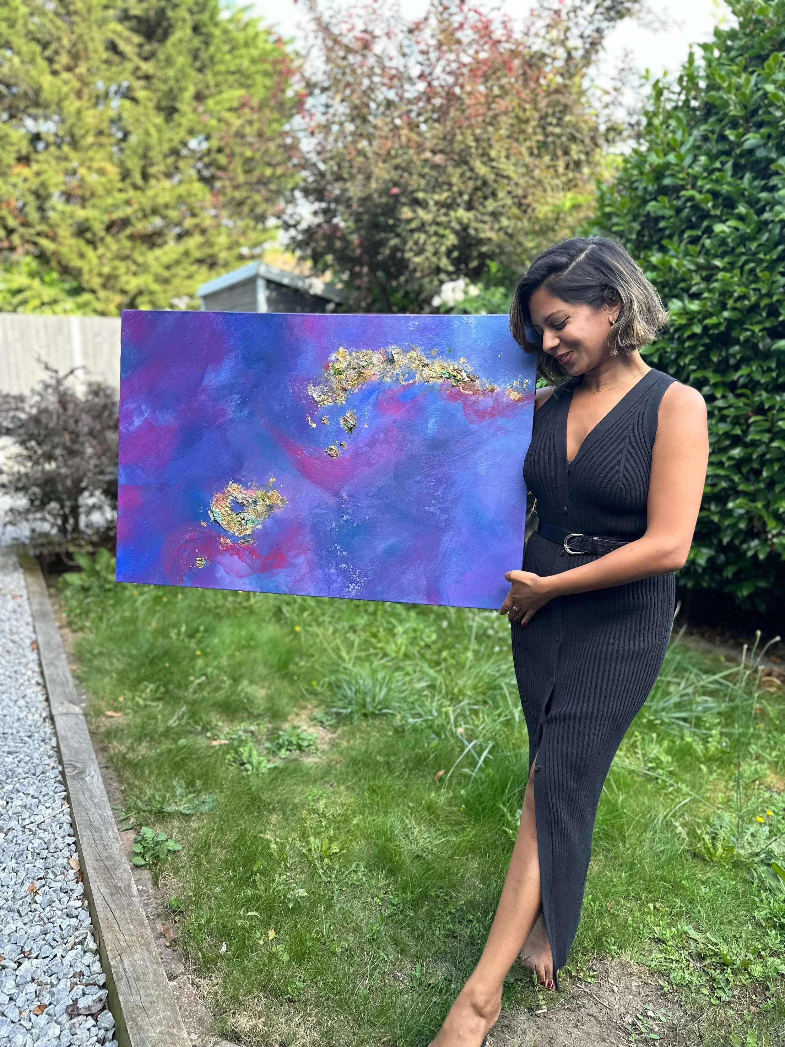 A woman in a black dress holding a colorful abstract painting outdoors in a garden with green trees and bushes.