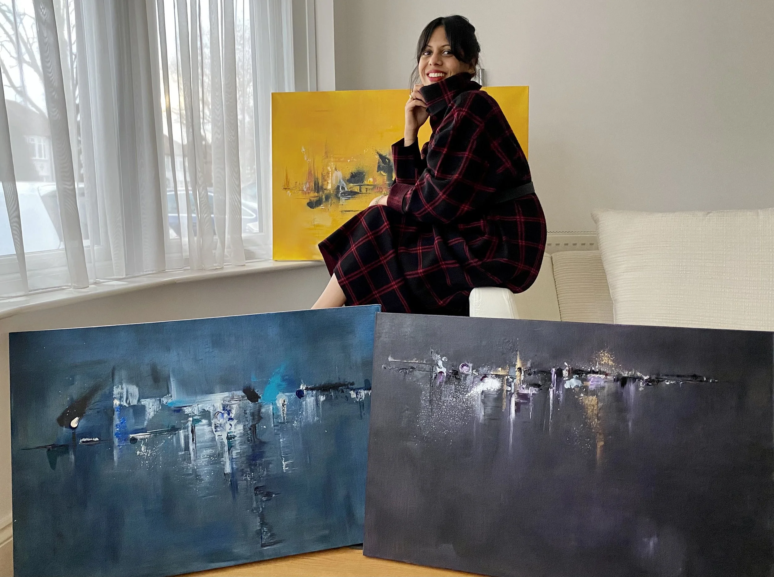 Young woman in a red and black plaid dress sitting on a white stool in front of abstract paintings on a table and a colorful painting on the window ledge, smiling at the camera.