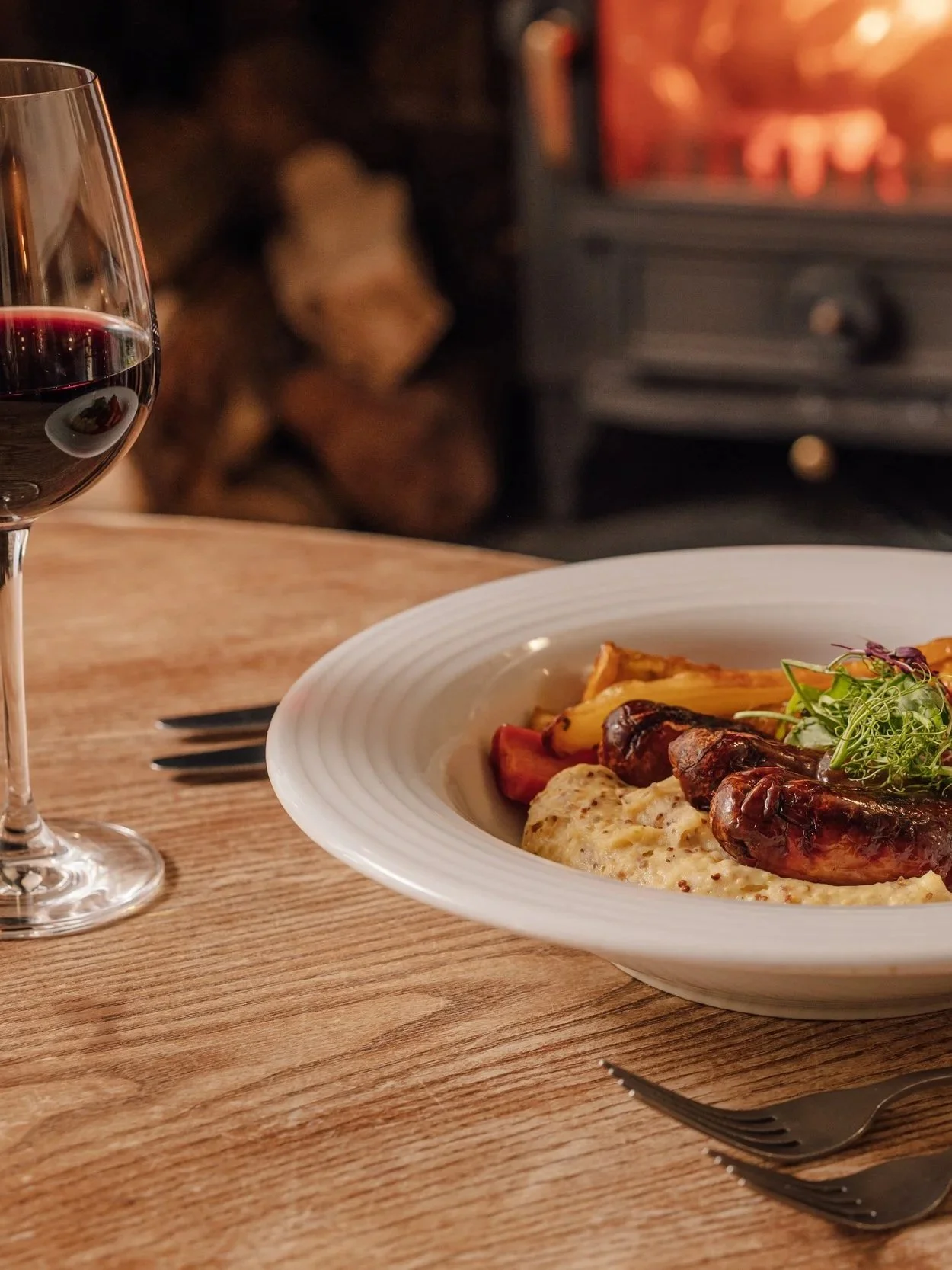 A white bowl of food on a wooden table, with roasted vegetables and possibly mashed potatoes with gravy, garnished with microgreens, beside a glass of red wine, in front of a fireplace with stacked logs and a glowing fire.