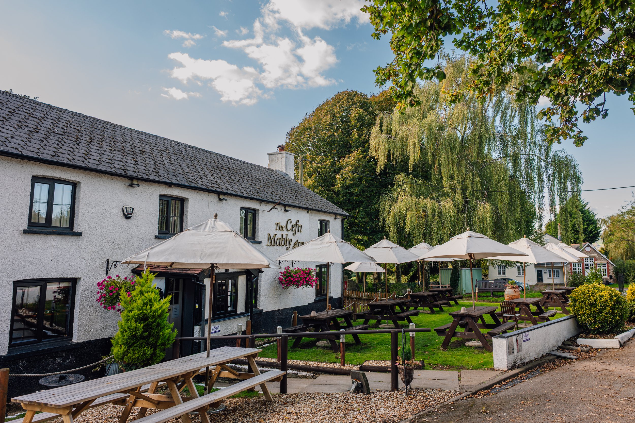 Exterior view of a pub with outdoor seating and umbrellas, surrounded by trees and plants, on a sunny day.