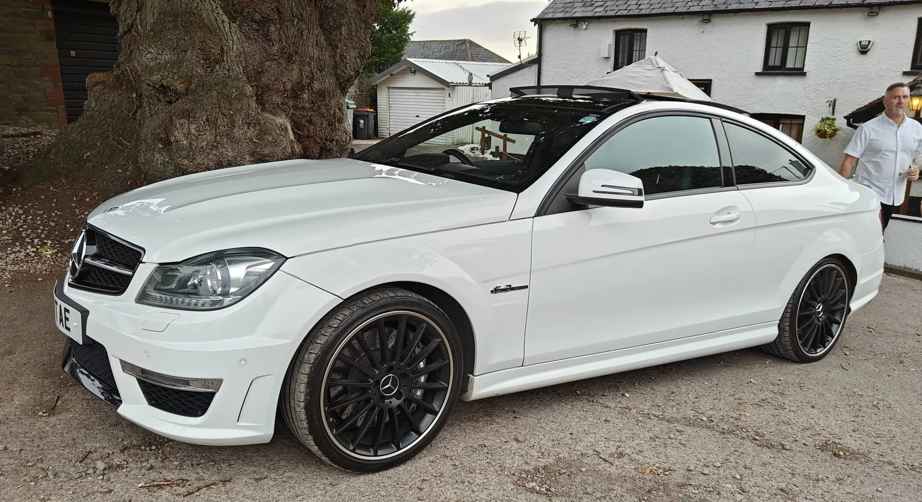 A white Mercedes-Benz coupe parked on a gravel driveway next to a large tree, with a man in a white shirt standing nearby.