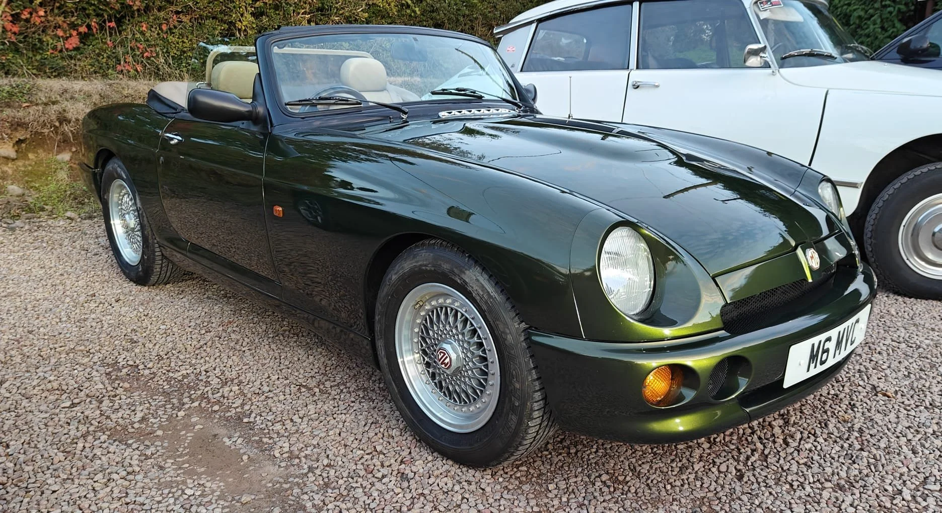 A black MG sports convertible car parked on gravel, with another white vintage car behind it, surrounded by trees and foliage.