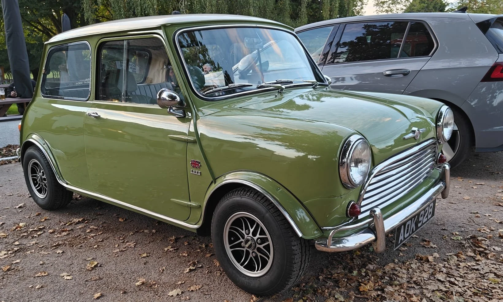 A vintage green Mini Cooper with a UK flag decal on the front side panel, parked on a gravel surface with fallen leaves, next to a modern gray car, surrounded by trees.