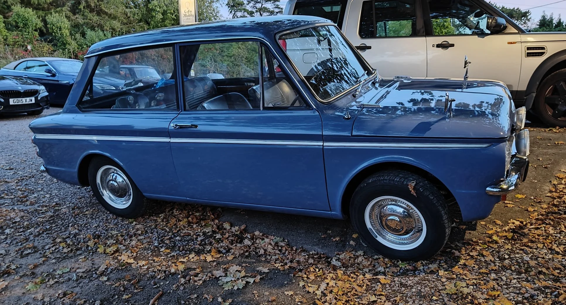 A vintage blue car with a two-tone paint job and chrome hubcaps parked on a gravel lot surrounded by modern cars and trees.