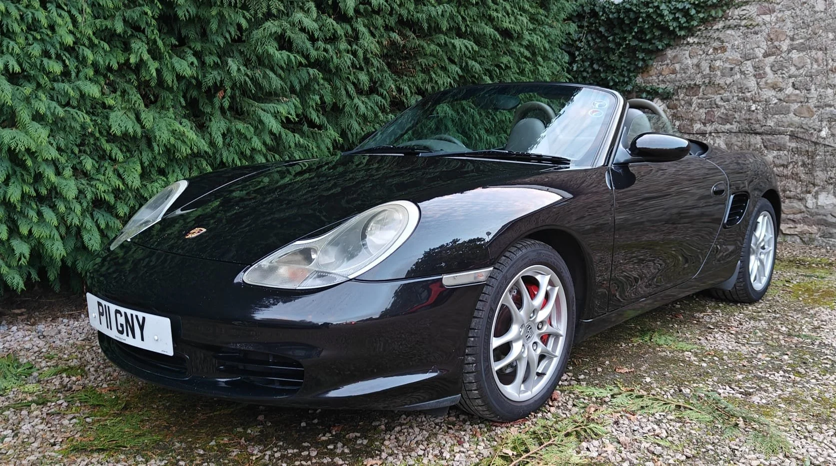 Black Porsche convertible car parked on gravel ground near green bushes and a stone wall.