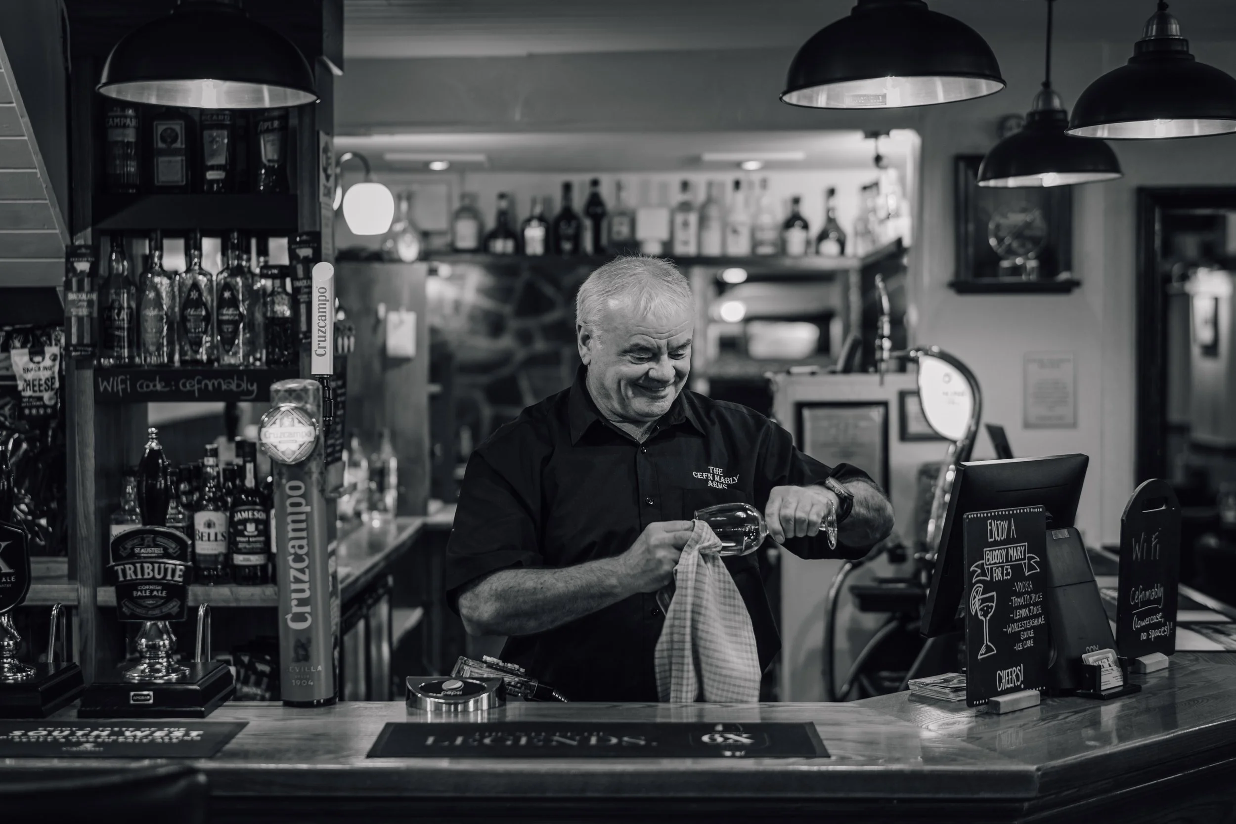 An older man behind a bar counter, smiling as he pours a drink, with bottles of alcohol on shelves in the background.