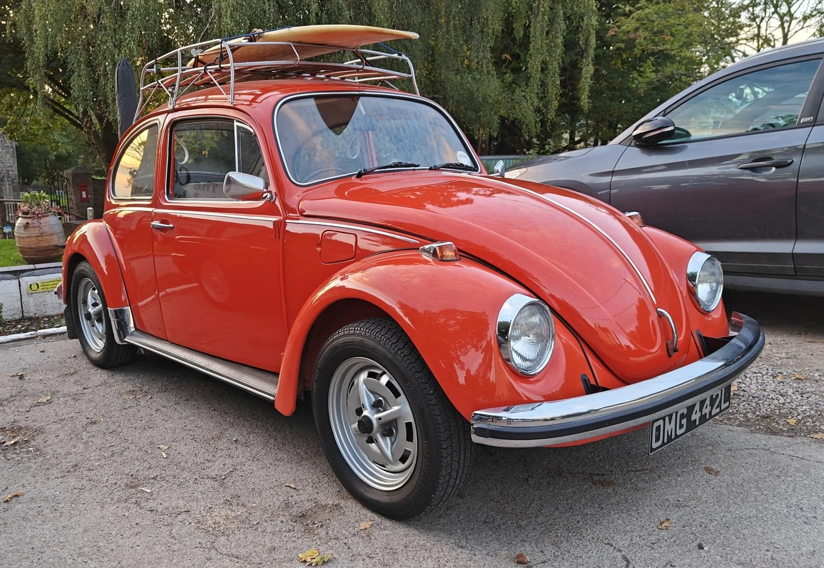 Red vintage Volkswagen Beetle with a surfboard and a kayak on the roof rack, parked on a street with trees in the background.