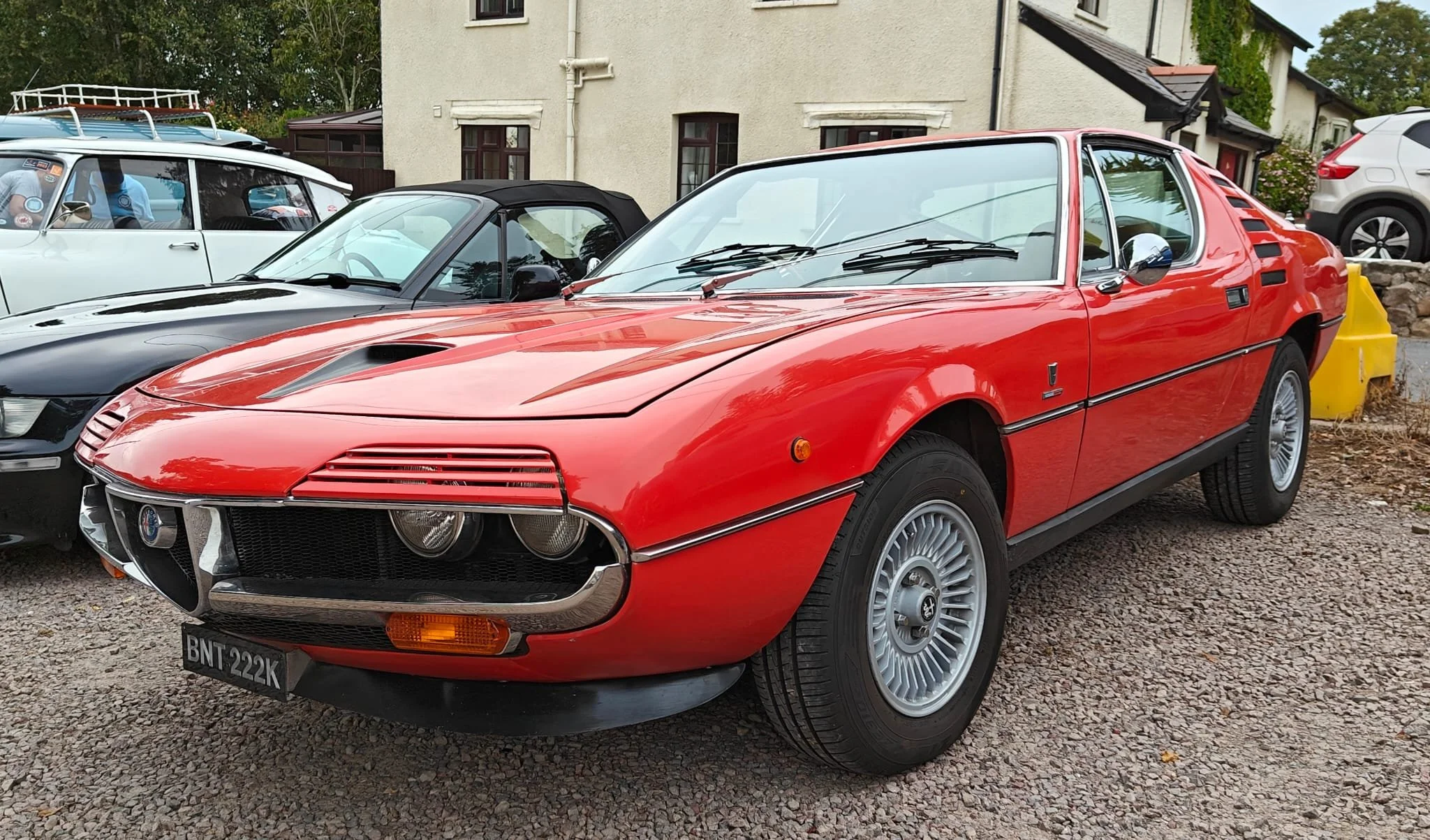 Red vintage sports car parked on gravel, surrounded by other classic cars, with a house in the background.