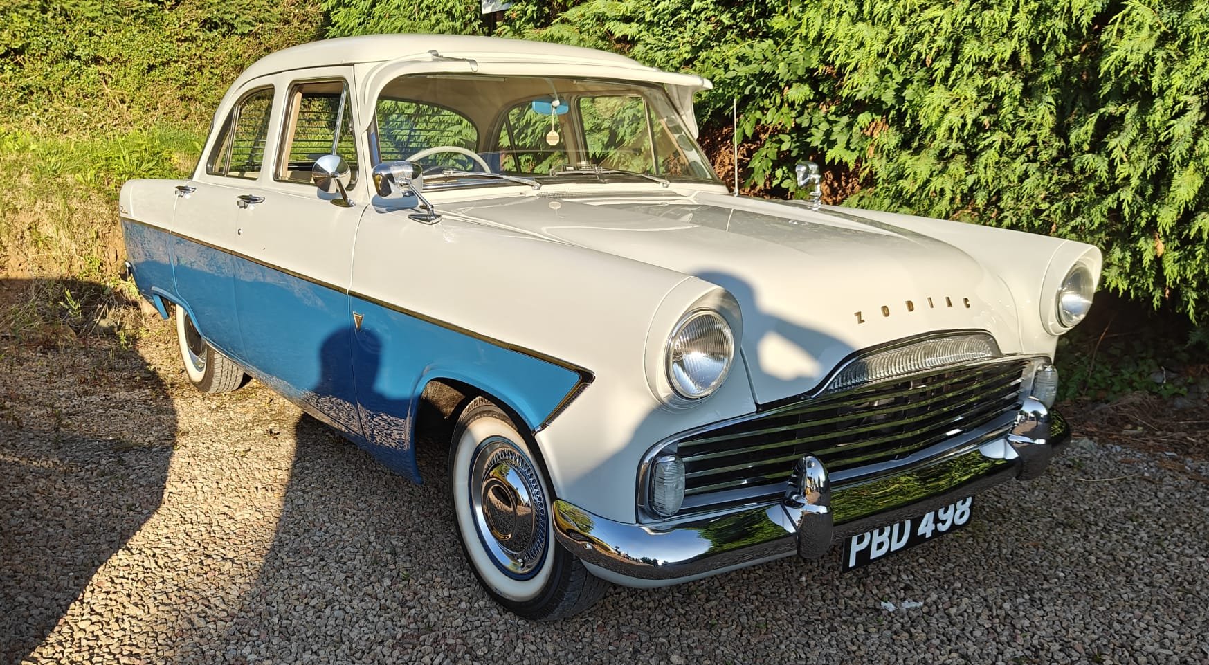 A vintage white and blue Zodiac car with chrome bumpers, grille, hubcaps, and side mirrors parked on gravel, with green bushes in the background.