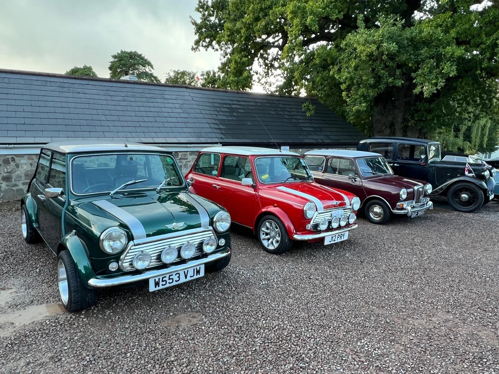 A row of vintage mini cars parked on a gravel surface with a stone building and green trees in the background.