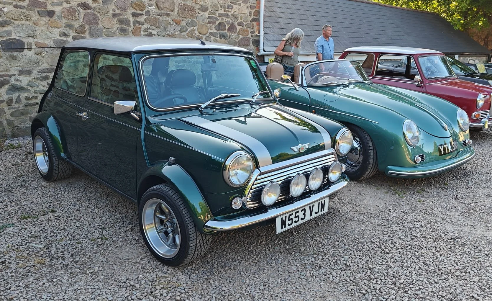 A classic Mini Cooper car with a dark green body and silver racing stripes, parked on a gravel surface, with other vintage cars and people in the background.