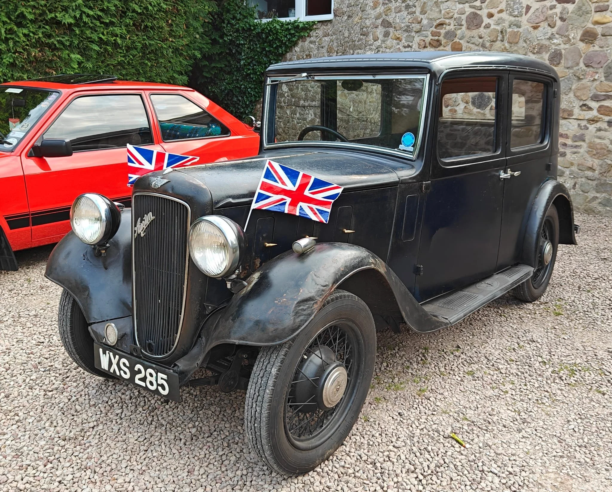 Vintage black car decorated with Union Jack flags parked on gravel, with a red sports car behind it. Stone wall with green foliage in the background.