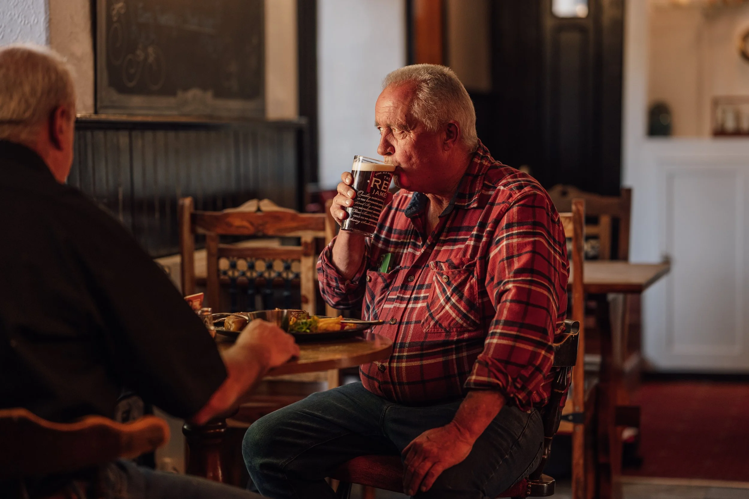 Older man in a red plaid shirt drinking a pint of dark beer while sitting at a wooden table in a cozy pub.