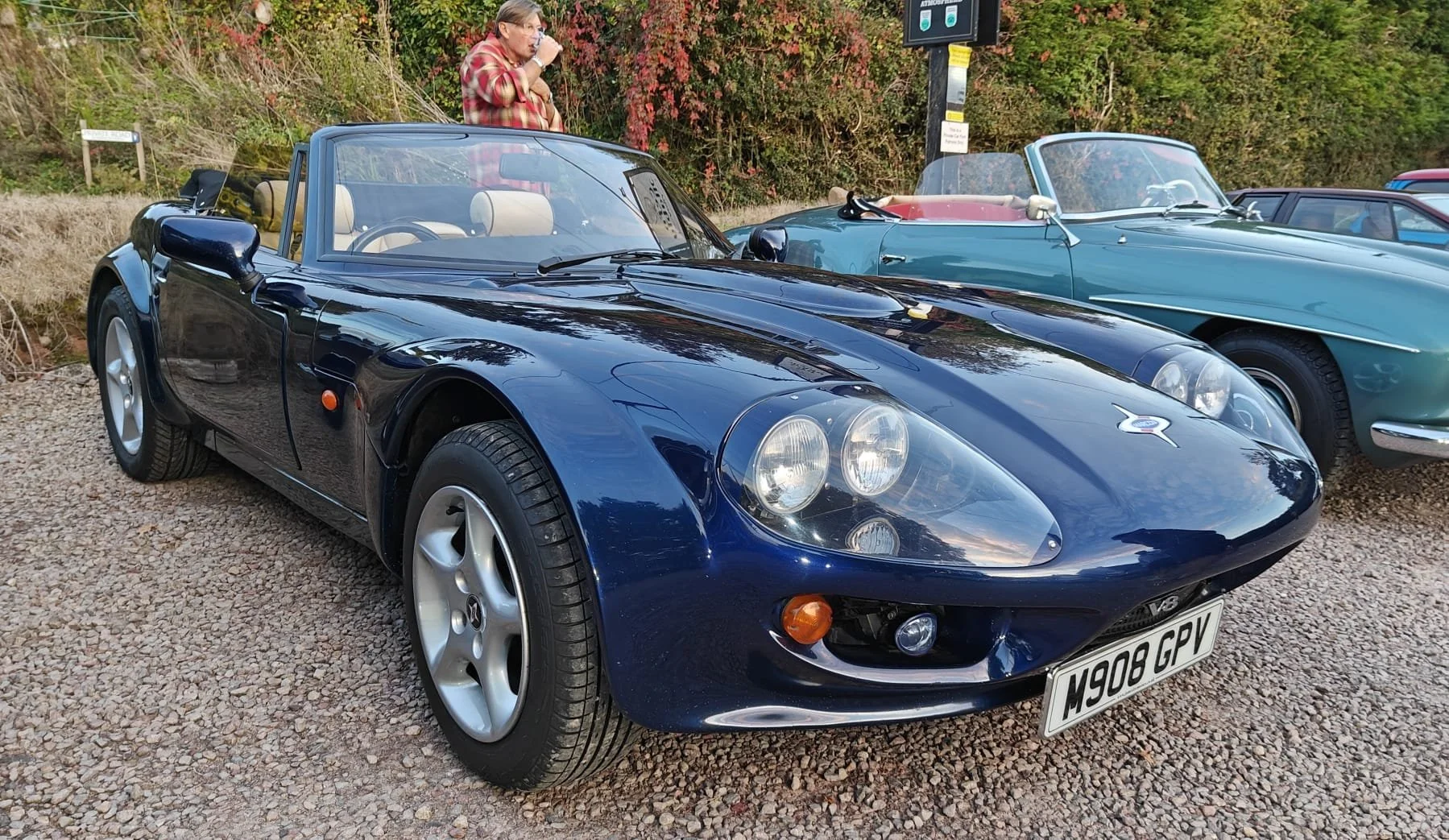 A black vintage convertible sports car with a beige interior parked on a gravel surface, with another vintage light blue convertible car beside it and autumn foliage in the background.