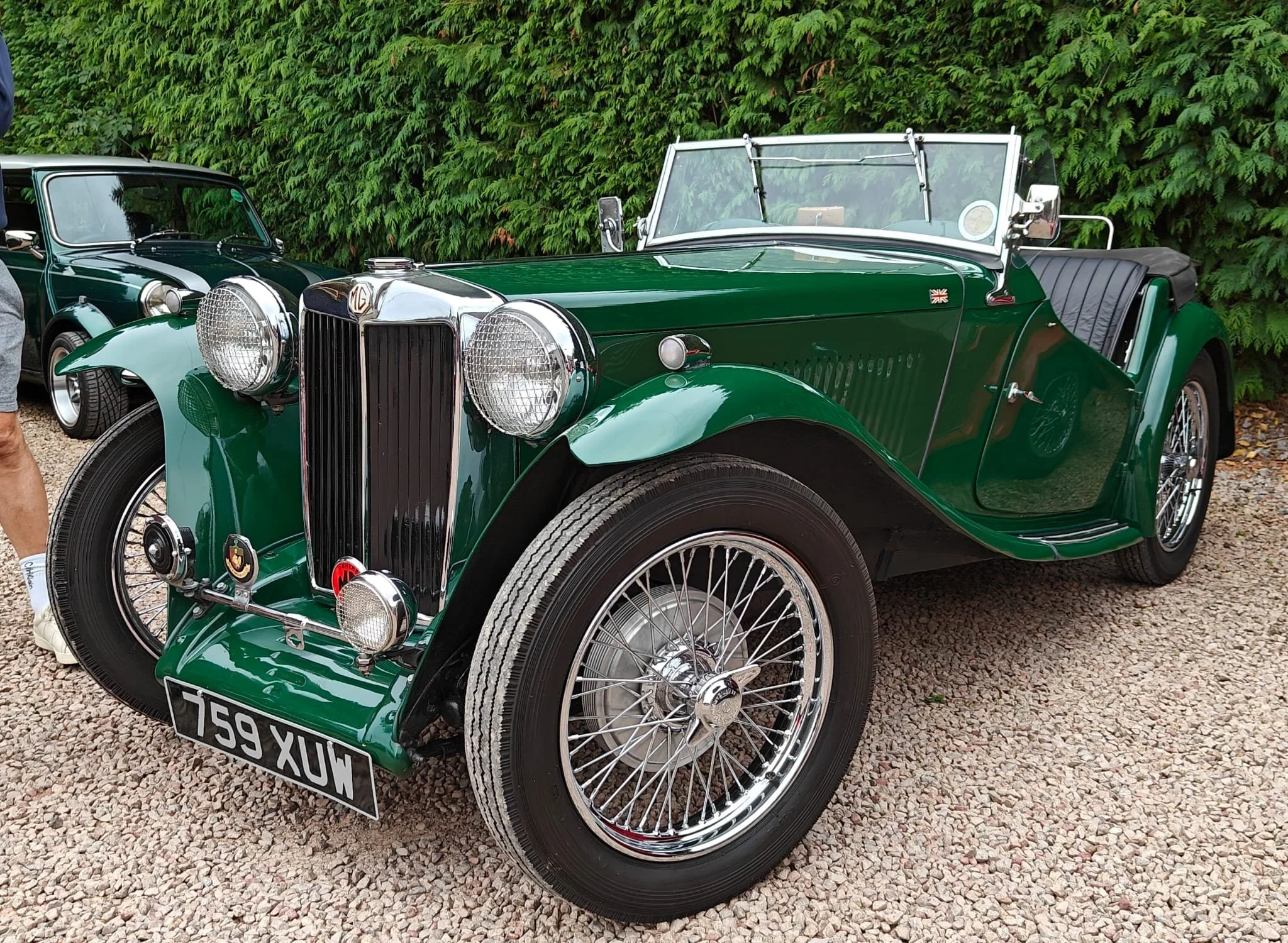 A vintage green MG classic car with chrome details and wire wheels parked on a gravel surface, with another classic car in the background and a green hedge behind.