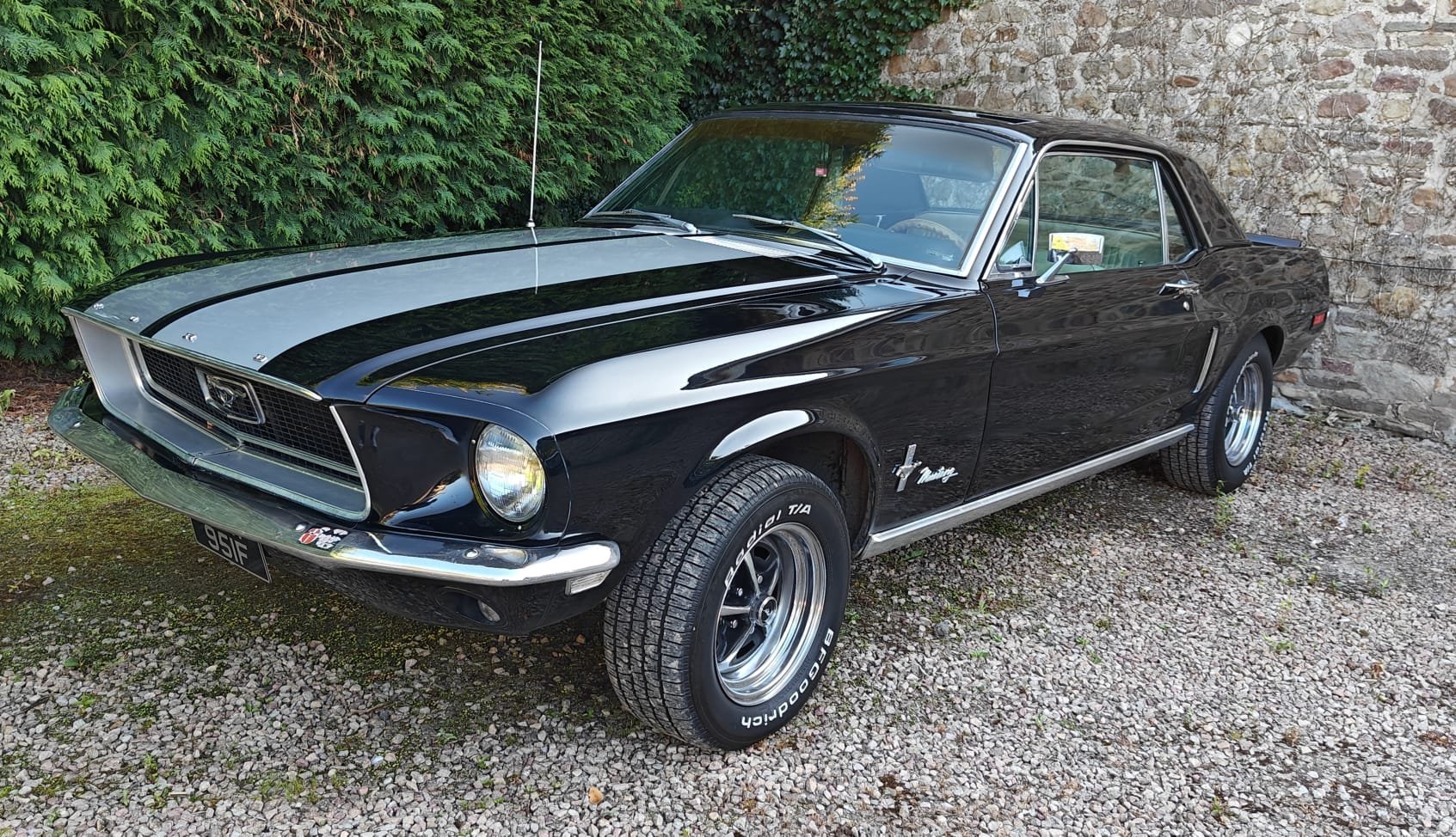 A classic black 1969 Ford Mustang with a white racing stripe parked on gravel next to a stone wall and green bushes.
