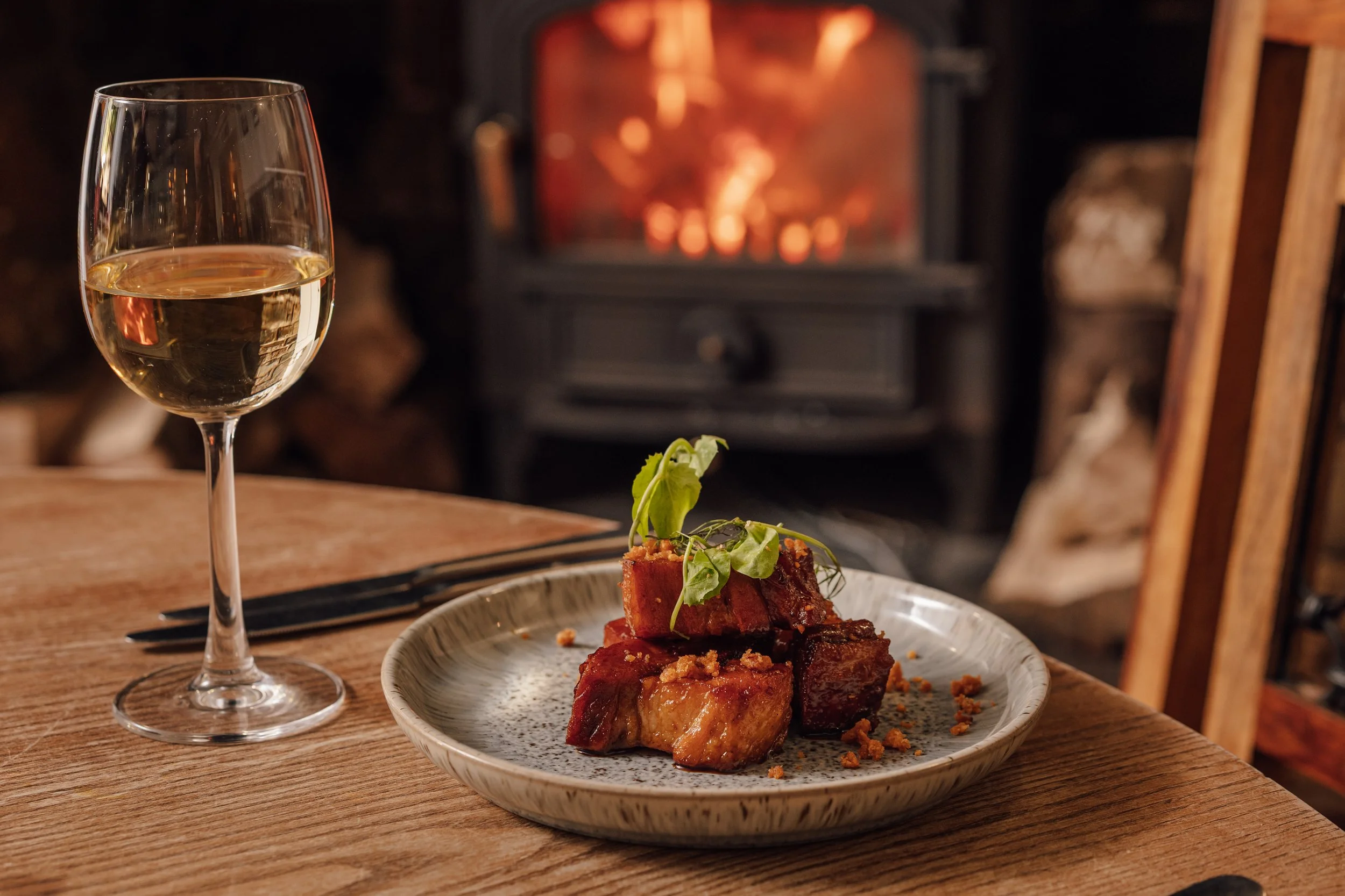 Glass of white wine and a plate of pork belly with garnish on a wooden table in front of a fireplace.